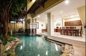 A swimming pool with a stone border, a wooden deck, and a dining area with chairs and a table. - PB - 7BR - V5 & V1 7BR Private Pool Villa, Walk to Seminyak Beach (Seminyak)