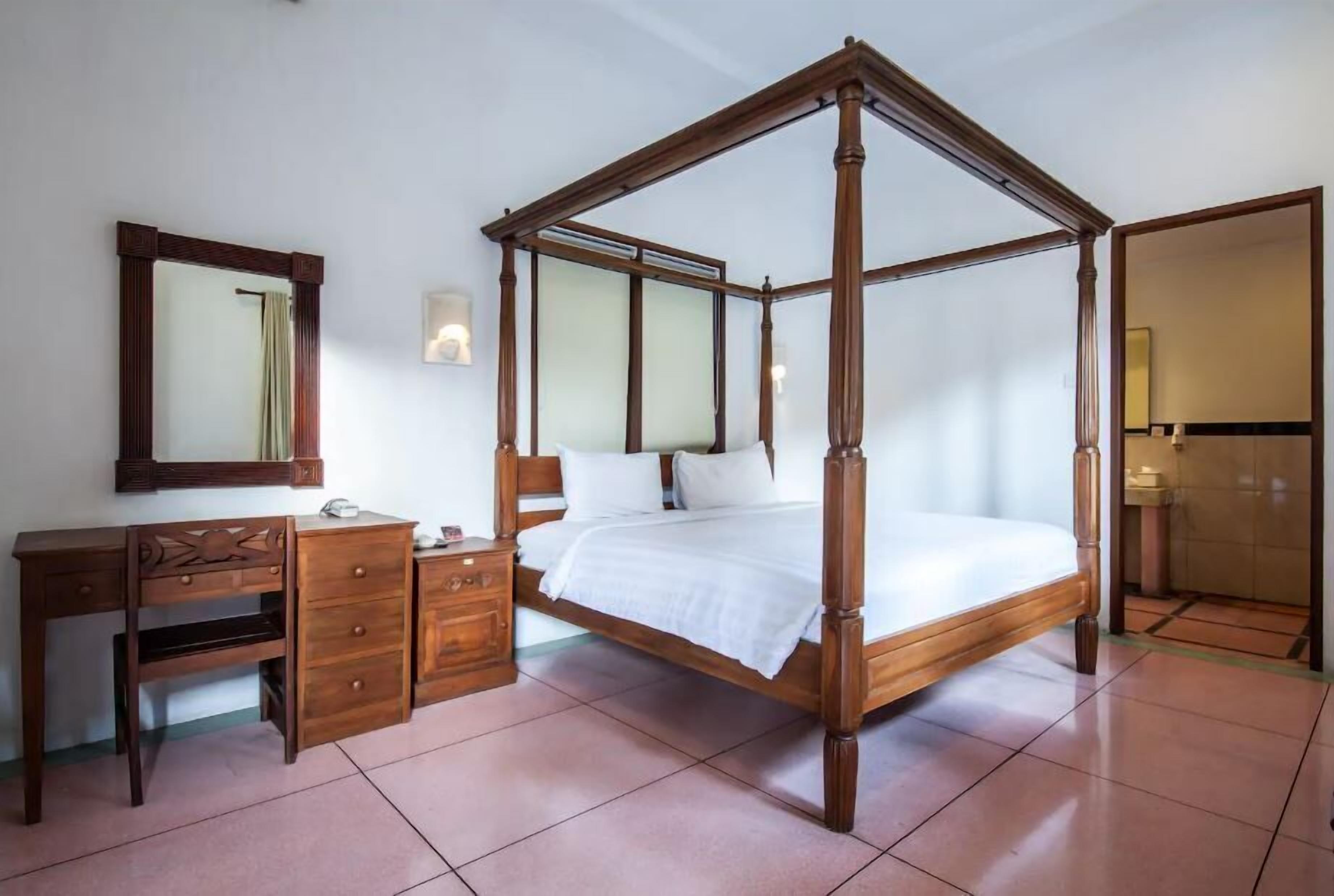 A four-poster bed with a wooden frame, a wooden desk with drawers, a mirror, and a wall-mounted light fixture.