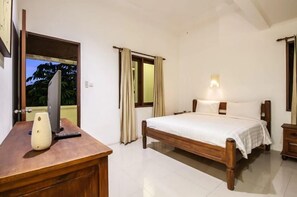 A bedroom with a wooden bed, a bedside table, a desk with a vase and remote control, and a window with curtains. - PB - 7BR - V5 & V1 7BR Private Pool Villa, Walk to Seminyak Beach (Seminyak)