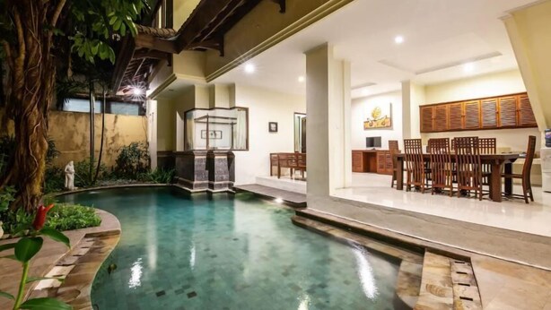 An indoor swimming pool with a wooden deck, a seating area with tables and chairs, and a small statue. - PB - Villa 5 - 4BR · 4-BR Private Pool Villa Walk to Seminyak Beach (Seminyak)