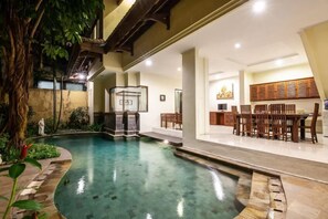Une piscine intérieure dotée d’une terrasse en bois, d’un espace détente avec des tables et des chaises, et d’une petite statue.