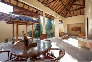 A spacious indoor area with a round glass table and wooden chairs, a thatched roof, and a view of greenery outside.
