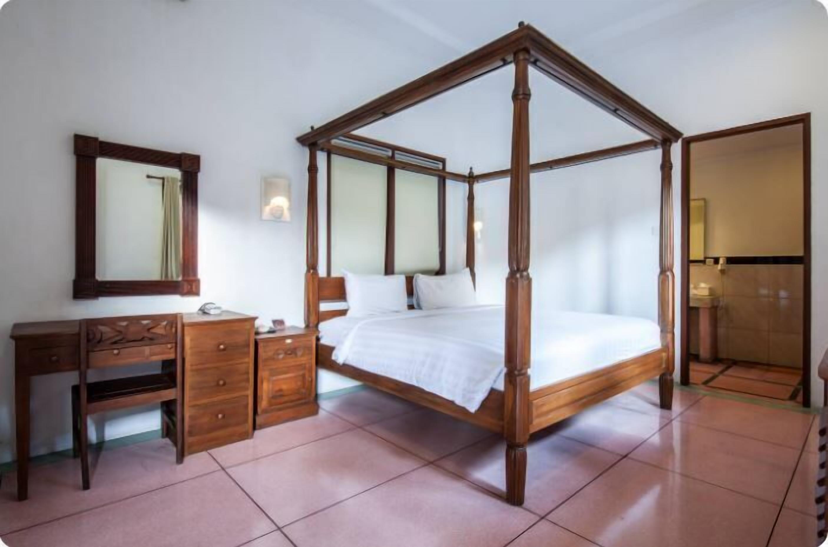 A four-poster bed with a wooden frame, a wooden desk with drawers, a mirror, and a wall-mounted light fixture.