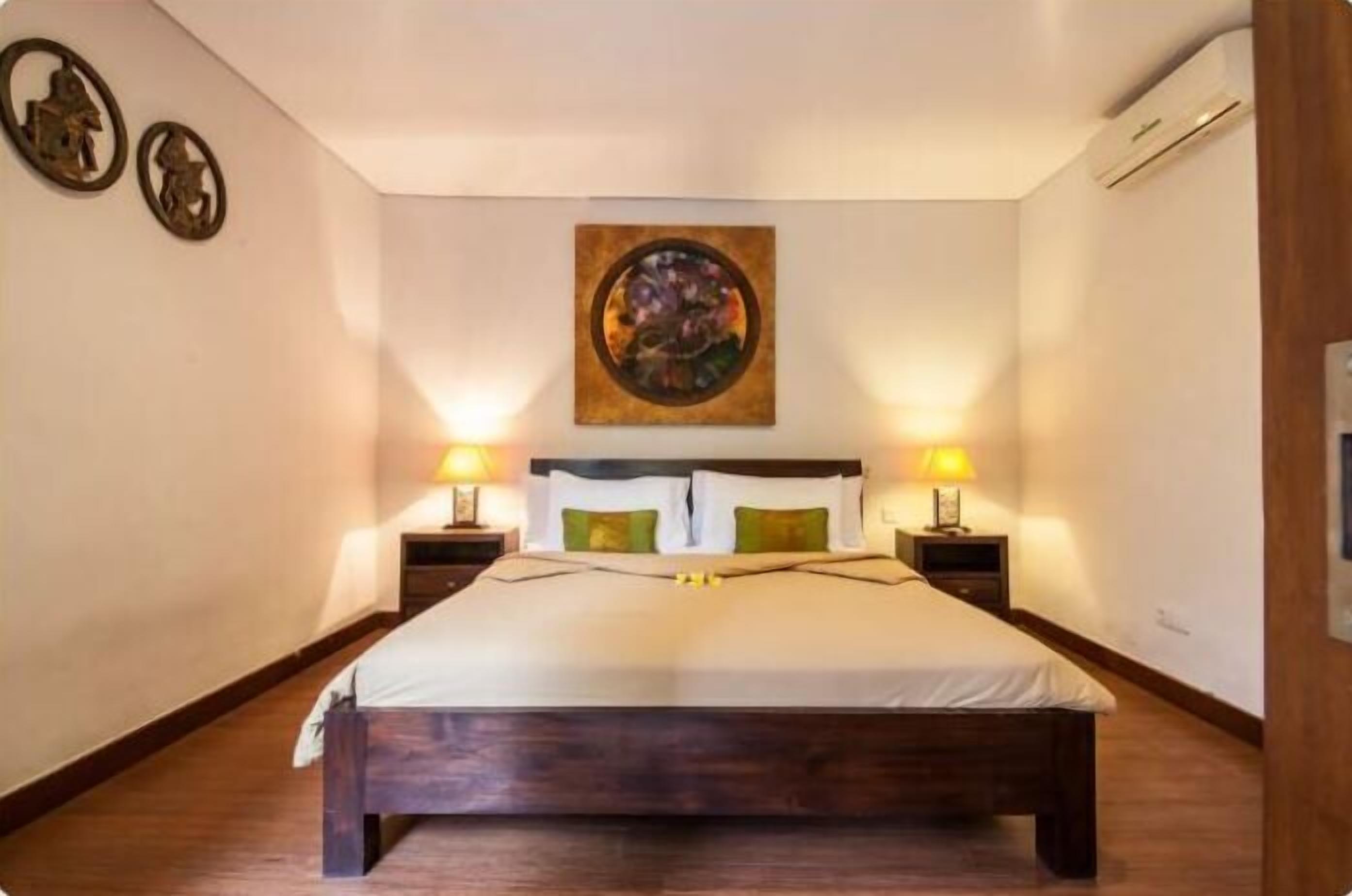 A bedroom with a wooden bed, two bedside tables with lamps, a wall-mounted painting, and a ceiling-mounted air conditioner.