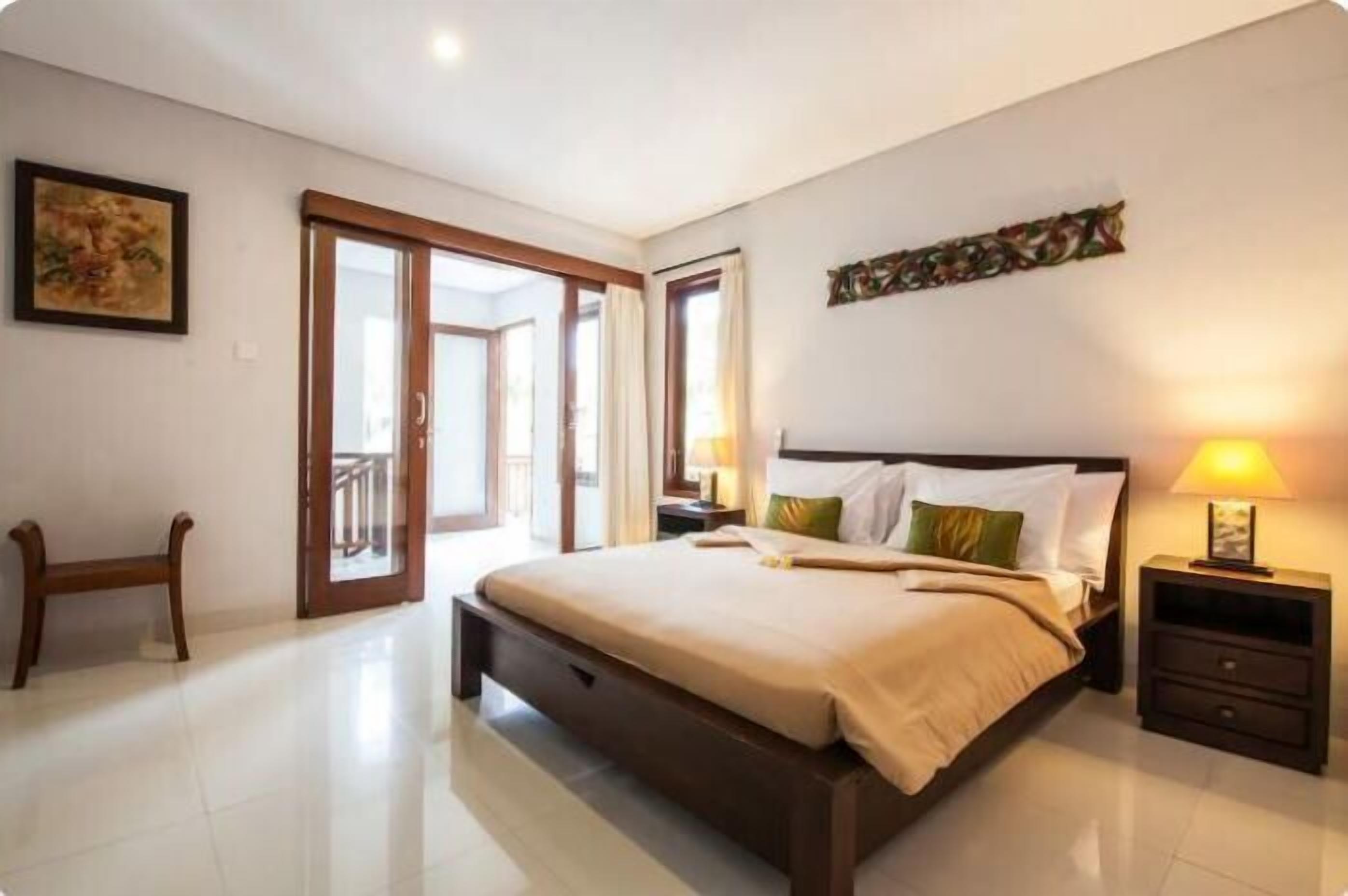 Quarto com cama grande, mesa de cabeceira, um banco e porta para uma varanda.