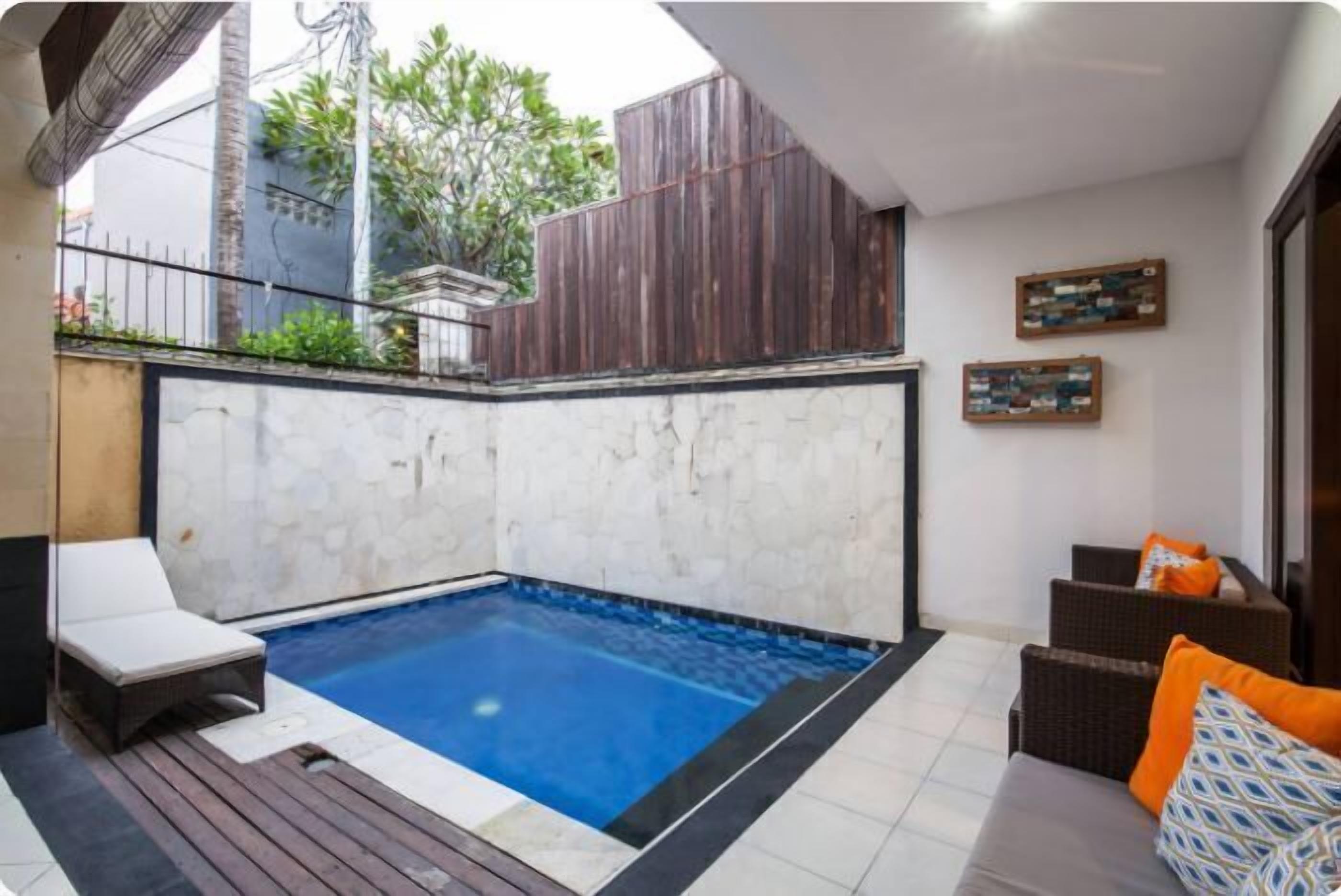 A swimming pool with a wooden deck, a white wall, and a seating area with cushions.