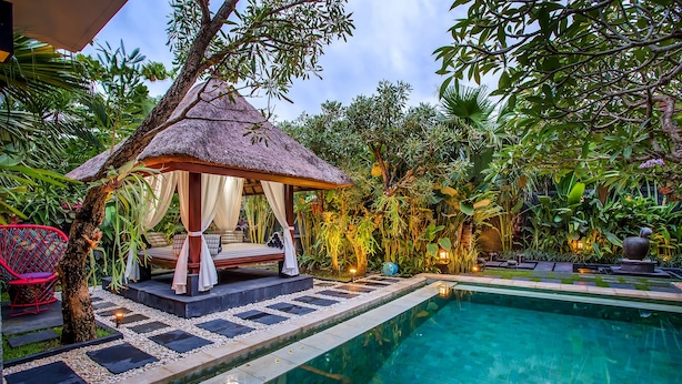 A thatched-roof gazebo with a cushioned seating area, a swimming pool, and a well-maintained garden with various plants. - Samudra · 3BR Luxury Private Pool Villa Bali (Kerobokan)