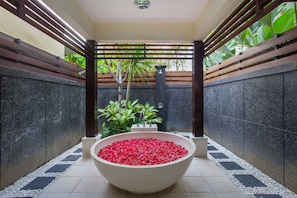 Un espace de détente avec un grand bassin en pierre rempli de pétales de roses, entouré d’un sol carrelé et d’une pergola en bois offrant une vue sur une végétation luxuriante.