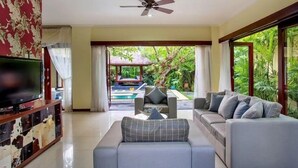A modern living room with a sofa, TV, and a ceiling fan. A pool and outdoor seating are visible through the glass doors. - Samudra · 6BR Luxury Family Pool Villa Umalas Bali (Kerobokan)