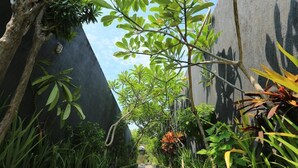 A narrow pathway flanked by lush greenery and a dark wall, leading to a building in the distance.
