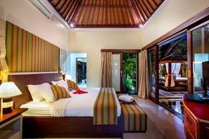 Een ruime slaapkamer met een groot bed, een nachtkastje met lamp, een bankje en uitzicht op een weelderige tuin via een schuifdeur van glas.