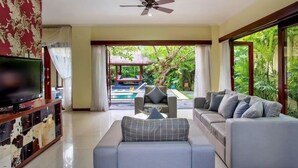A living room with a TV, sofas, and a ceiling fan. There is a view of a pool and greenery outside. - Samudra - 1 · 1BR Luxury Private Pool Villa Bali (Kerobokan)