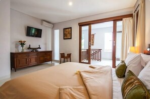 Quarto de hotel com uma cama grande, televisão, escrivaninha com um vaso de flores e varanda com guarda-corpo.