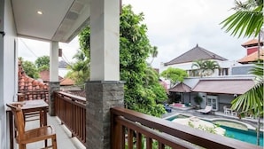 A balcony with a wooden table and chairs, overlooking a swimming pool and a garden area with various plants and a building in the background. - SMV - garden - 8-BR · 8BR Garden Villa Walk to Beach and Shops Legian (Legian)