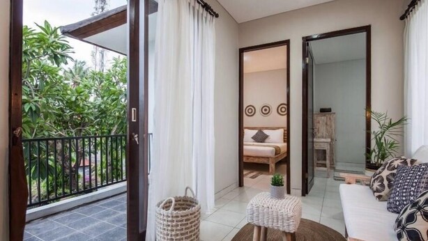 Ein Balkon mit Blick auf Grünflächen, ein Korbwicker-Korb, ein kleiner Hocker und eine Couch mit gemusterten Kissen.