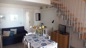 A living room with a sofa, a dining table with a vase of flowers, a TV on a wooden cabinet, and a staircase.