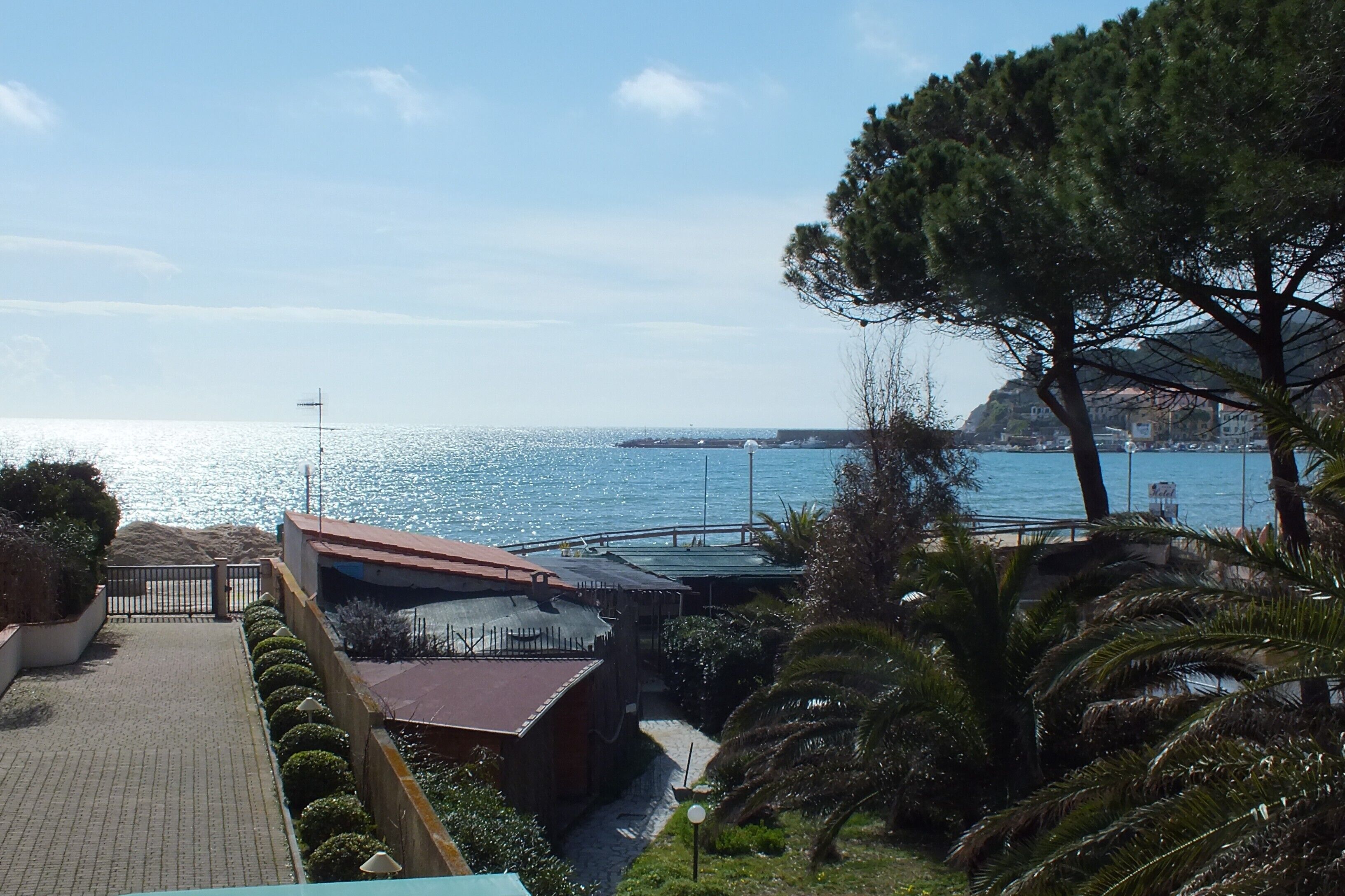 A coastal view with a paved walkway, a building with a red roof, and a clear blue sea.