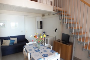 Un salon avec un canapé, une table à manger recouverte d’une nappe à motifs, un téléviseur posé sur un meuble en bois et un escalier.
