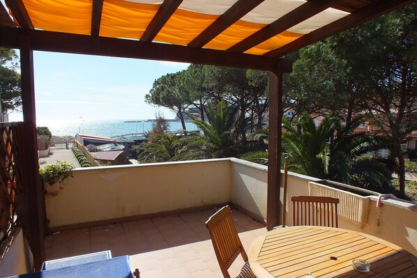 Un balcon avec une table et des chaises en bois, donnant sur une zone côtière bordée d’arbres et offrant une vue sur la mer.