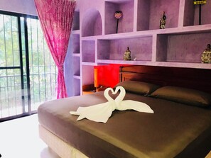 Un letto rifatto con cura, un asciugamano a forma di cuore, una lampada rossa e uno scaffale con oggetti decorativi.