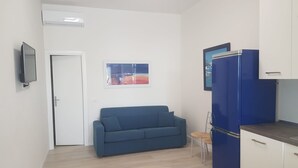 Un espacio habitable compacto con kitchenette, un sofá azul, una pequeña mesa y una puerta.