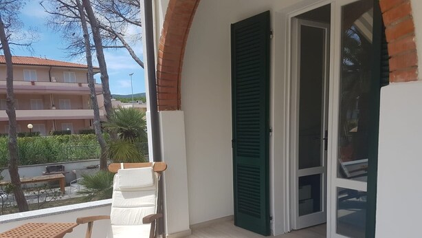 Un balcón con una silla de madera, una mesita pequeña y una ventana con contraventanas verdes.