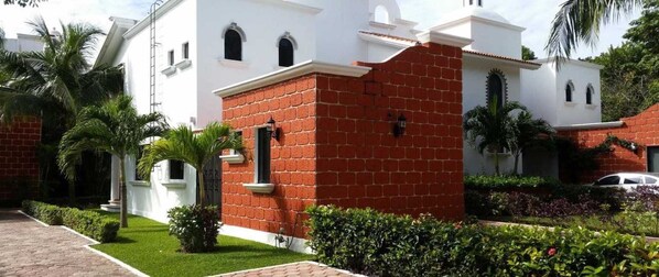 A building with a white facade and a red brick section, surrounded by well-maintained greenery and a paved walkway.