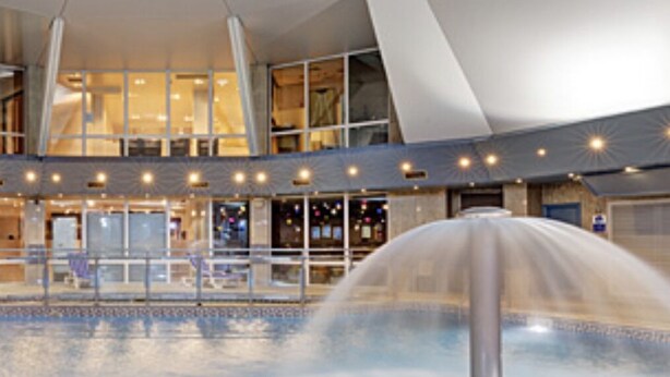 A large indoor swimming pool with a central fountain, surrounded by a glass enclosure and a modern architectural design. - Oakridge, St Mellion, Cornwall + 2 Leisure Passes (Saltash)