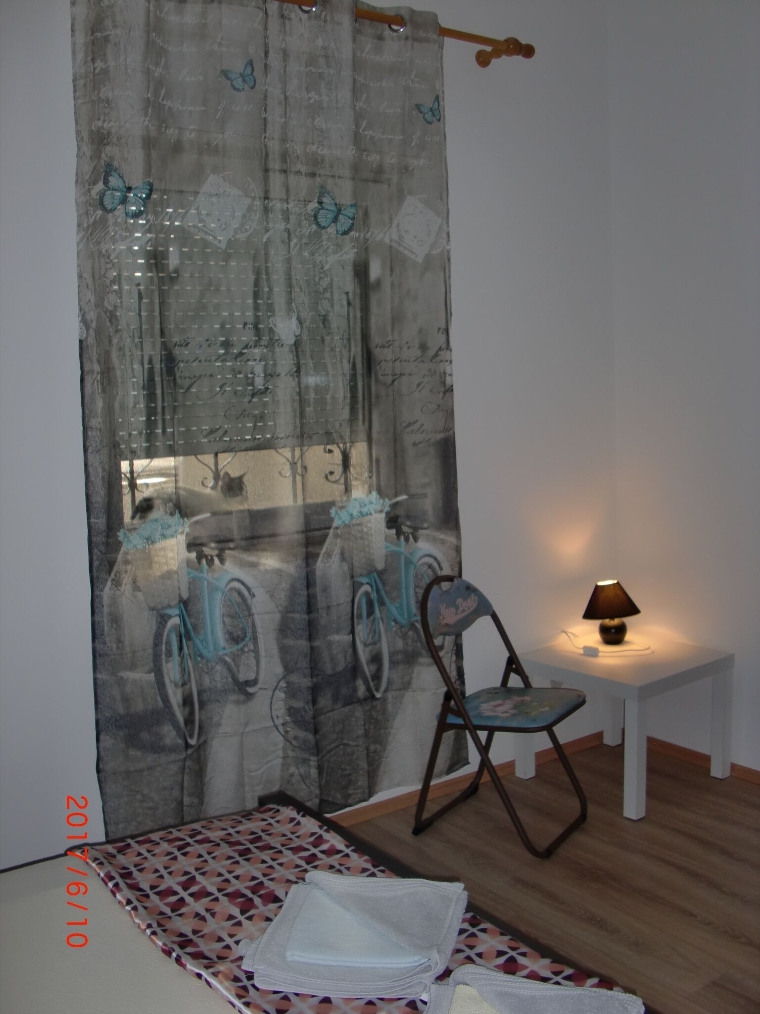 A room with a patterned curtain, a small table with a lamp, a chair, and a bed with a patterned bedspread.