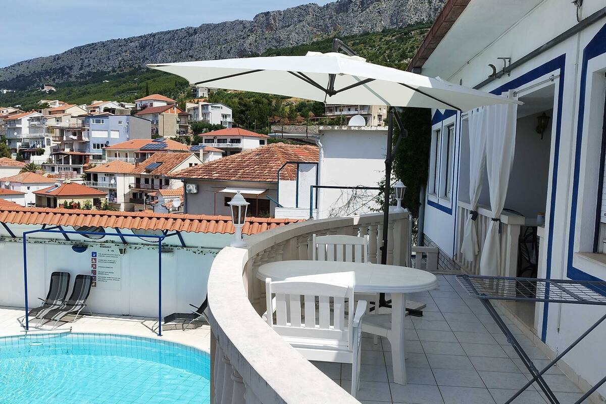 A poolside area with a white table and chairs, a white umbrella, and a view of a residential area with houses and mountains in the background.