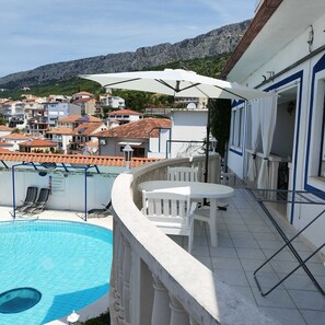 Un espace aménagé au bord de la piscine, avec une table et des chaises blanches, un parasol blanc, et une vue sur un quartier résidentiel avec des maisons et des montagnes en arrière-plan.