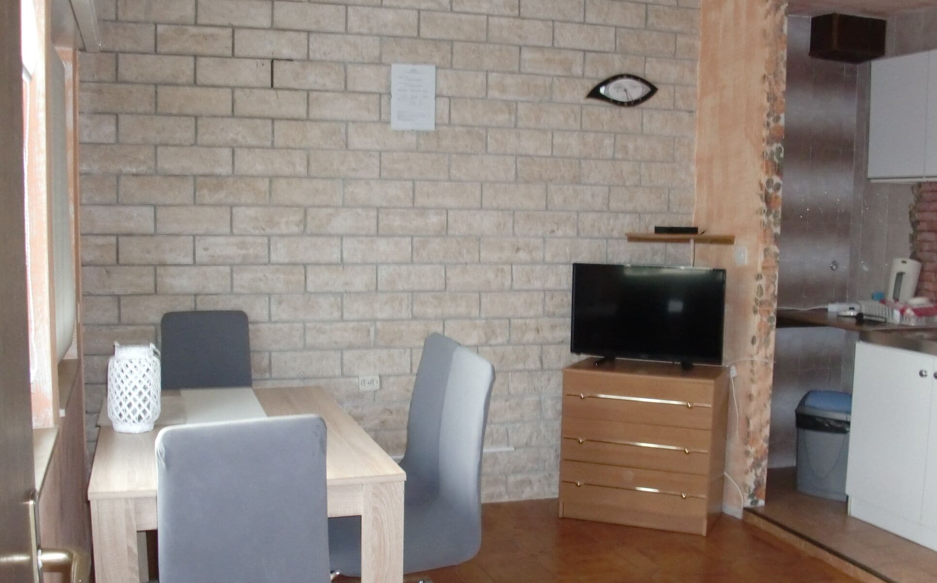 Een kleine keuken met een bakstenen muur, een houten kastje met een televisie, en een eethoek met een tafel en stoelen.