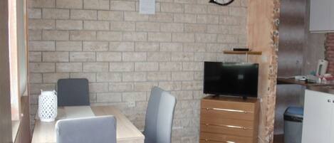 Een kleine keuken met een bakstenen muur, een houten kastje met een televisie, en een eethoek met een tafel en stoelen.