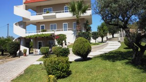 A multi-story building with a curved facade, a red-tiled roof, and a well-maintained garden with various shrubs and a palm tree. - Villa Olive Grove,superior Studio (Rhodes)