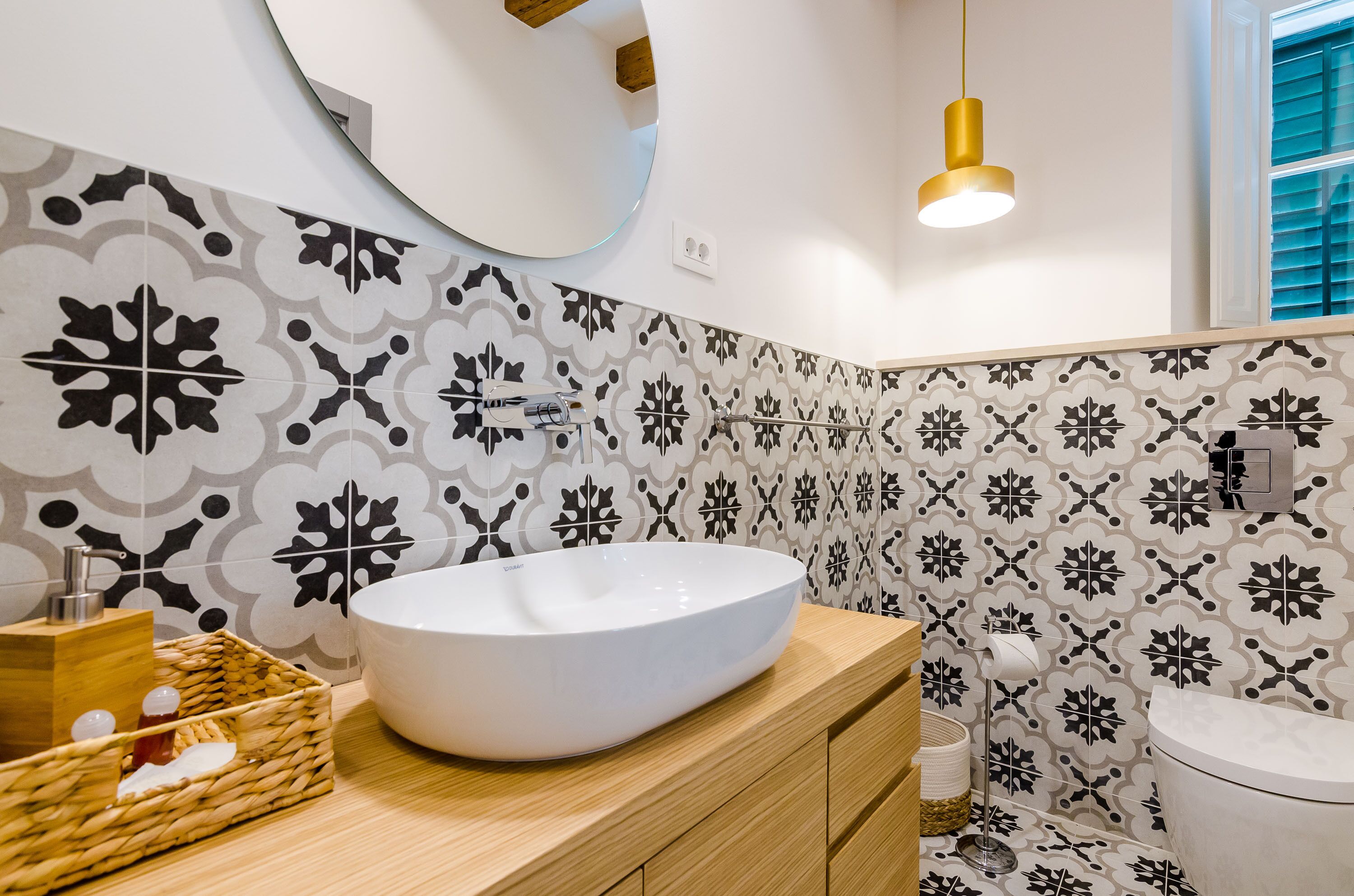 A modern bathroom with a large white vessel sink, patterned tiles, wooden cabinetry, a round mirror, and a pendant light.