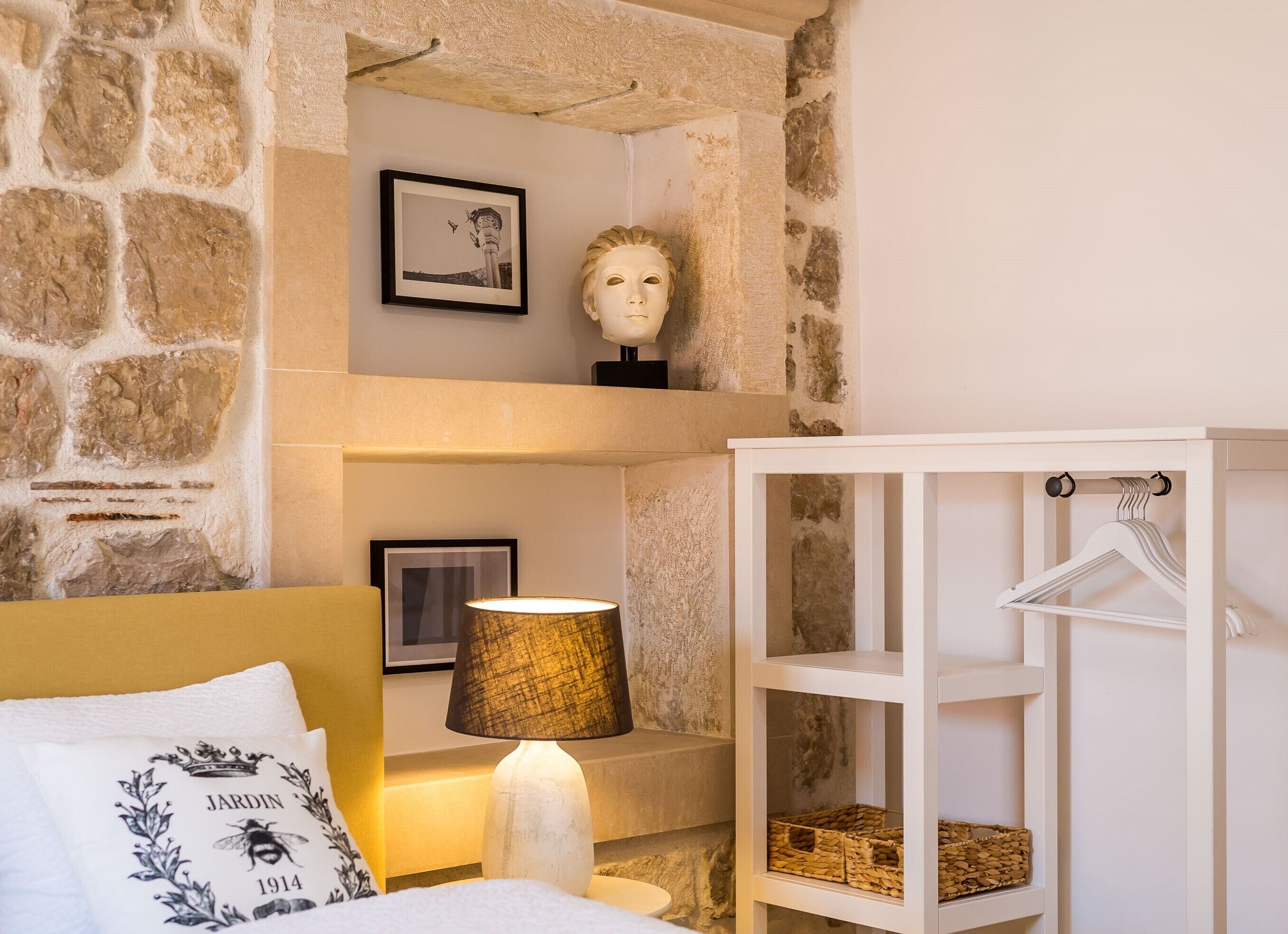A bedroom with a stone wall, a bed with a white headboard, a lamp, a framed picture, a bust sculpture, and a white cabinet with shelves.