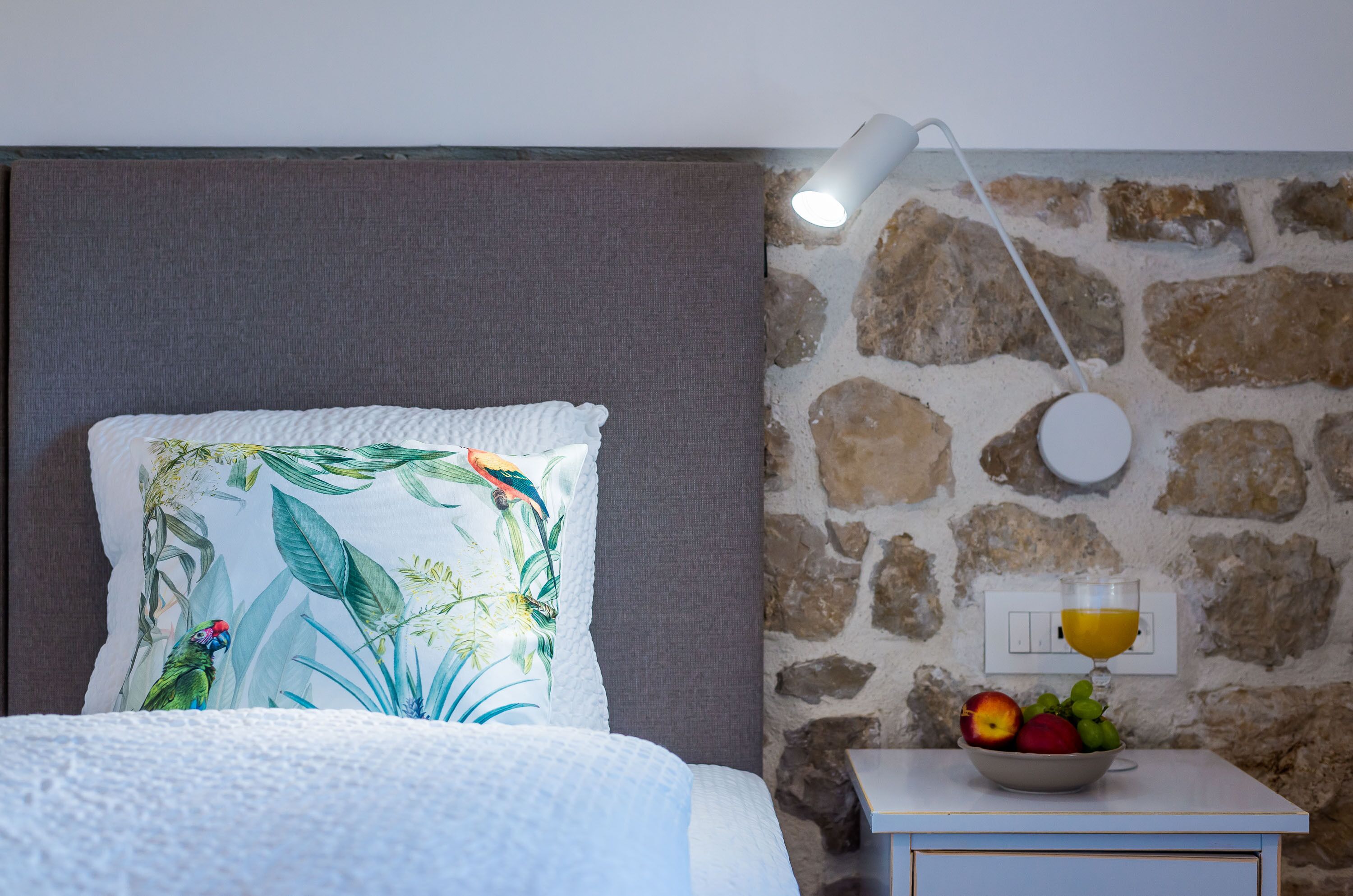 Une chambre à coucher avec un lit, une table de chevet sur laquelle se trouve un panier de fruits, une lampe fixée au mur et un mur en pierre.