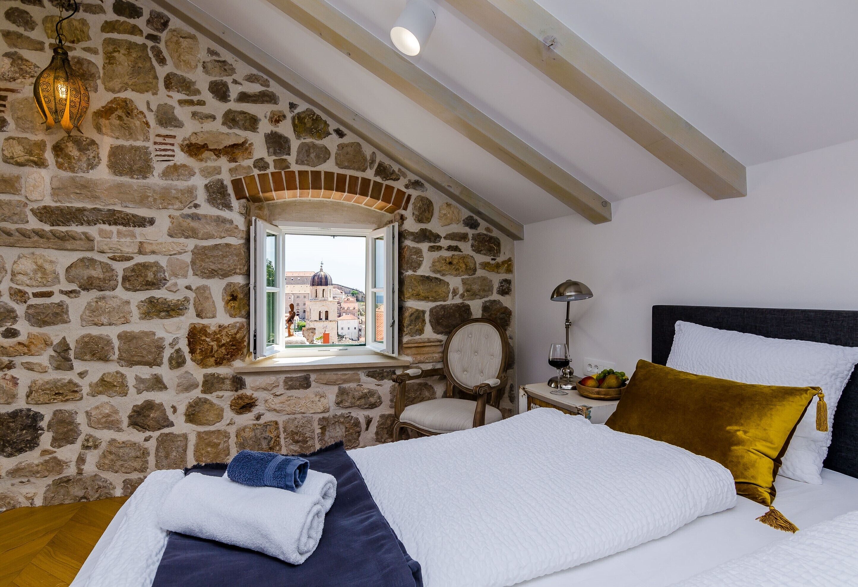 Quarto com parede de pedra, cama com roupa de cama branca e azul, cadeira de madeira e mesa de cabeceira com luminária e tigela de frutas.