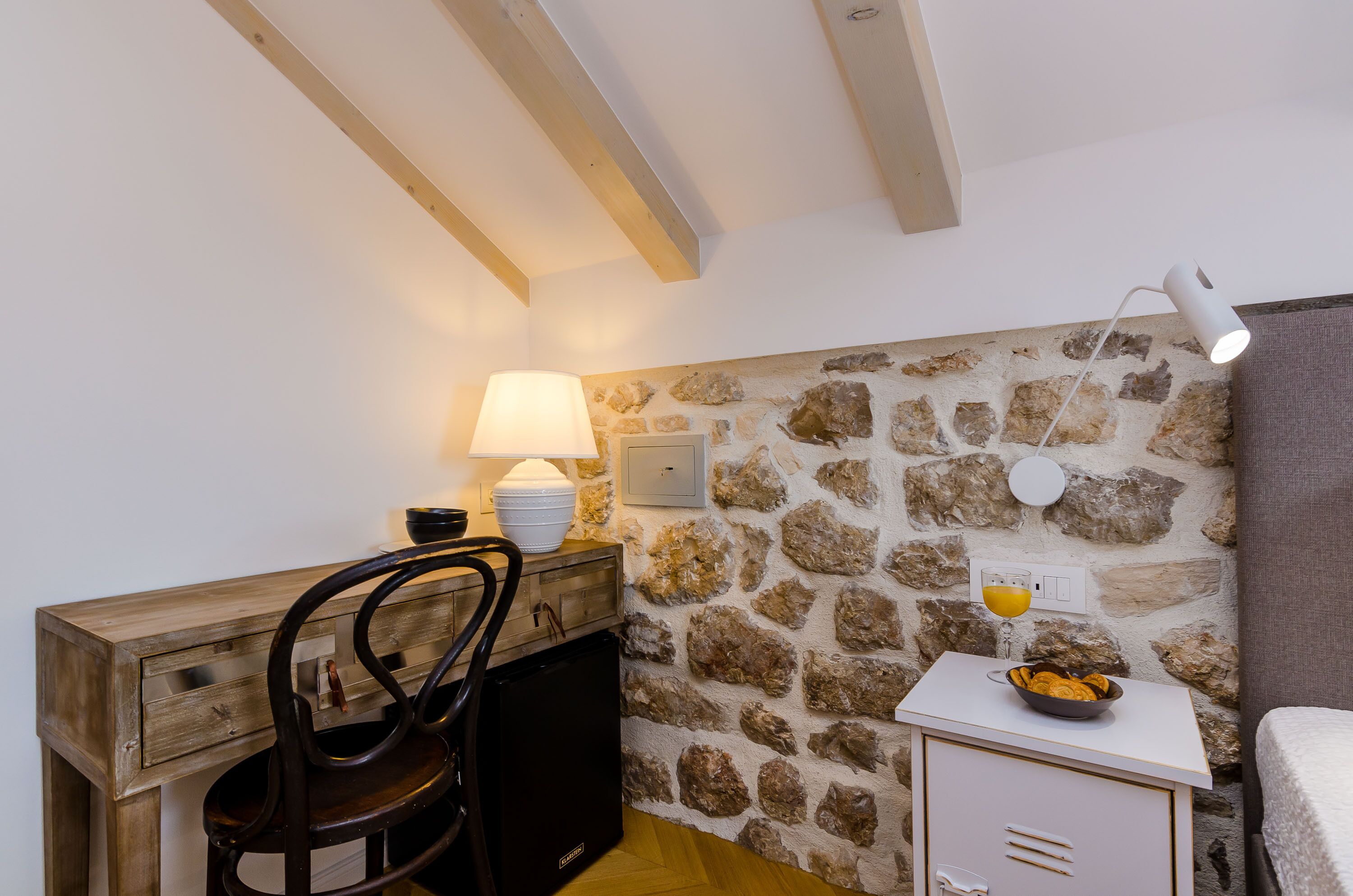 A compact room with a stone wall, a wooden desk with a chair, a small table with a bowl, a lamp, and a bed.