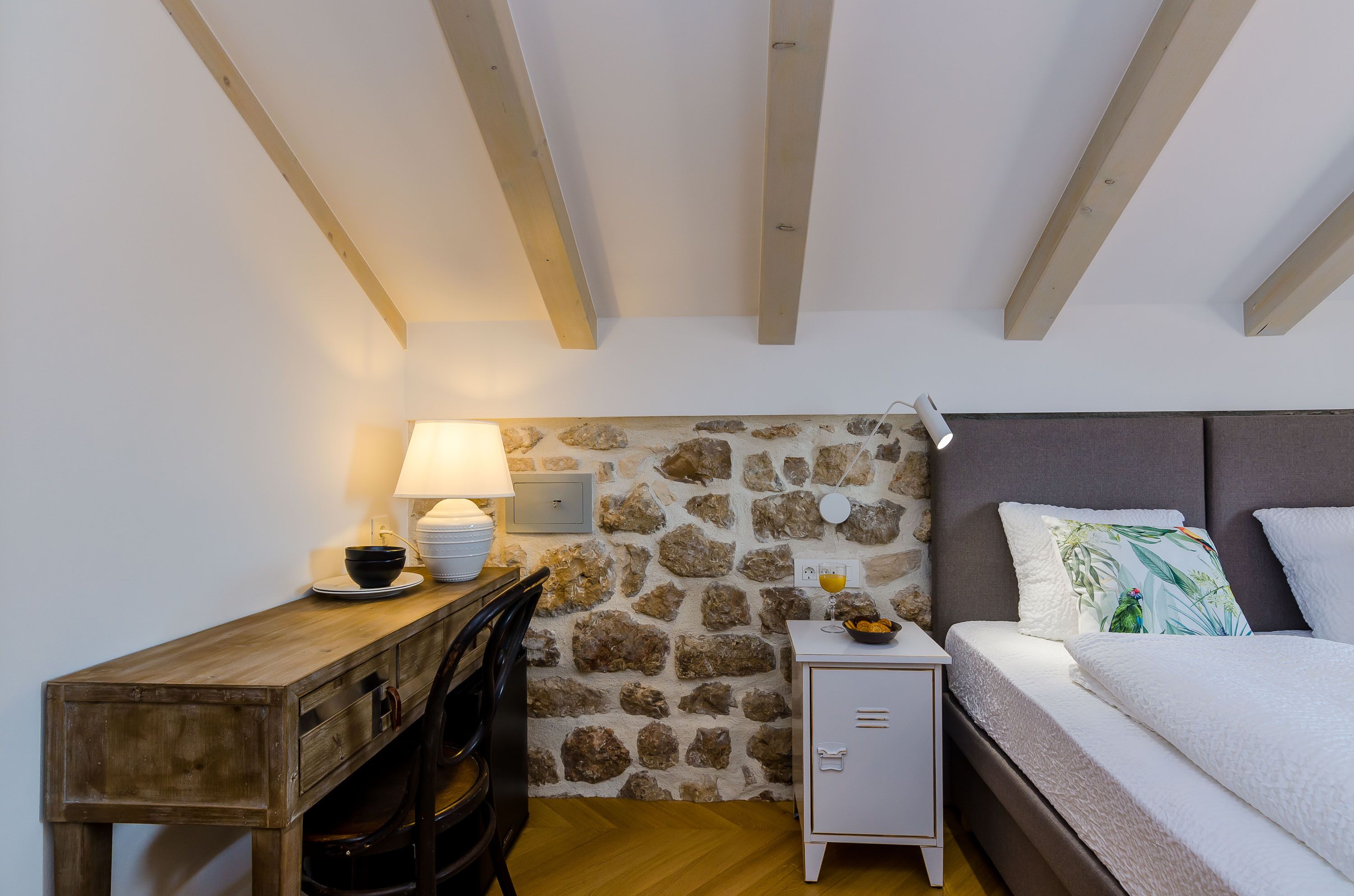 A bedroom with a stone wall, wooden beams, a bed with pillows, a bedside table with a lamp, and a small cabinet.