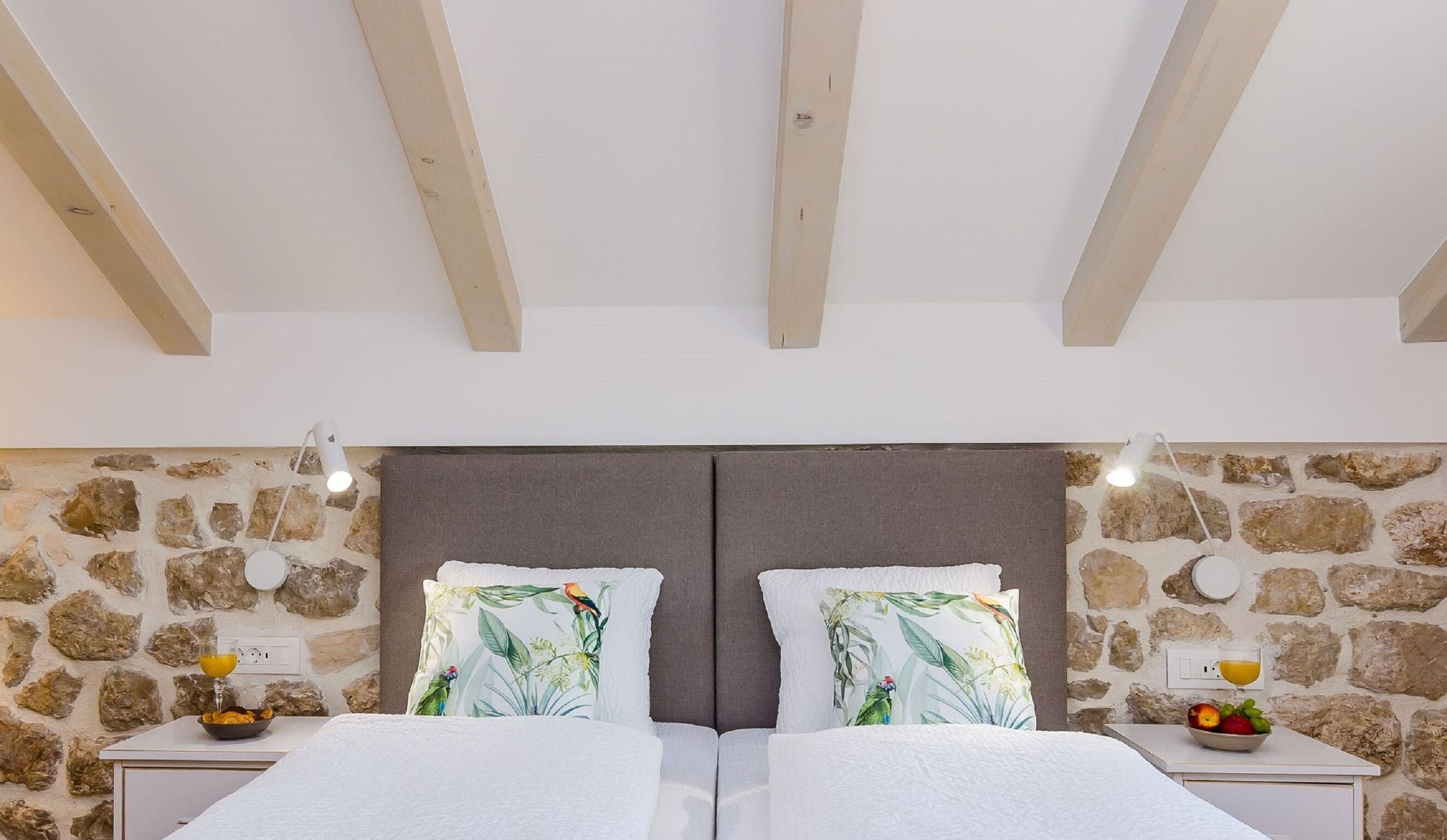 A double bed with a grey headboard, two nightstands with lamps, a stone wall, and a wooden ceiling.