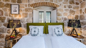 Un dormitorio con paredes de piedra, una cama con ropa de cama blanca, dos mesitas de noche con lámparas y una ventana con contraventanas.