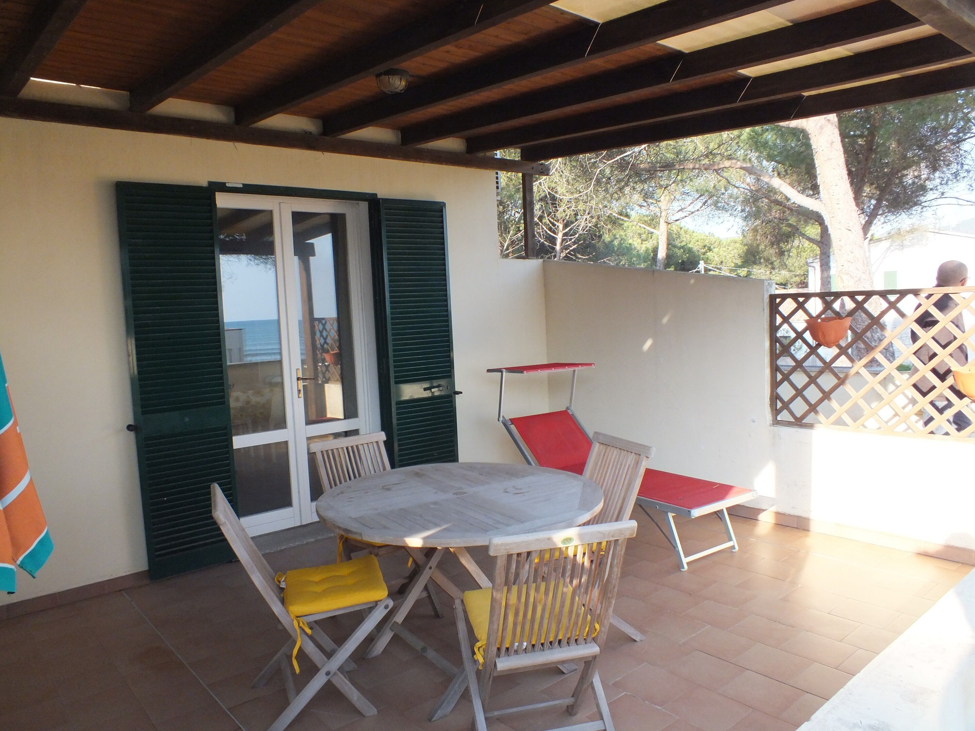 An outdoor patio with a round table and chairs, a view of the sea through a glass door, and a wooden ceiling.