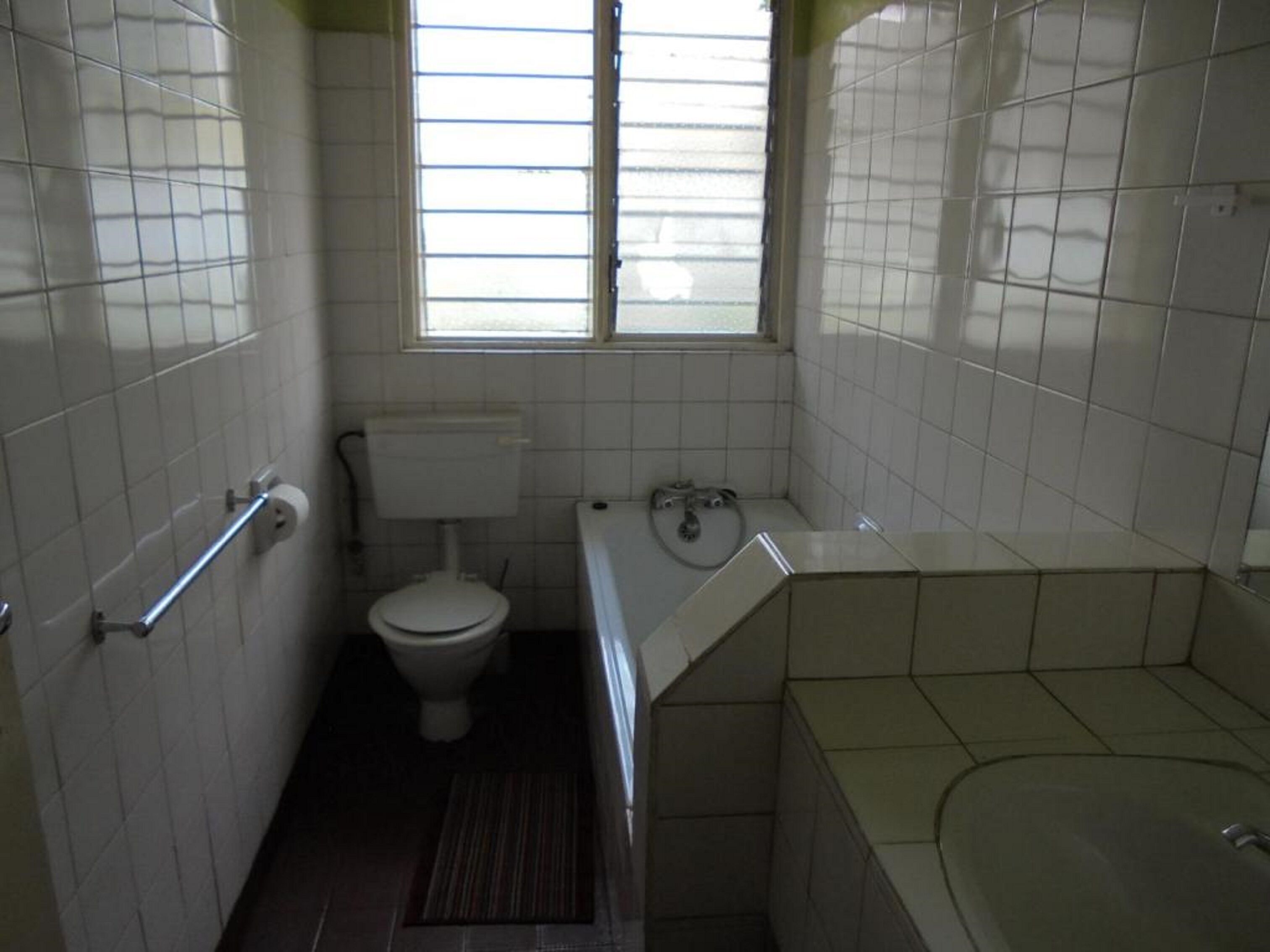 Un baño pequeño y estrecho con azulejos blancos, un inodoro, una bañera y una ventana con persianas.
