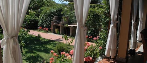 A balcony with white curtains, a garden with pink flowers, and a tiled floor.