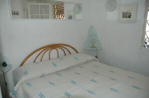 A bedroom with a wooden headboard, a bed with a white sheet featuring a blue pattern, a bedside lamp, a mirror, and a small window.