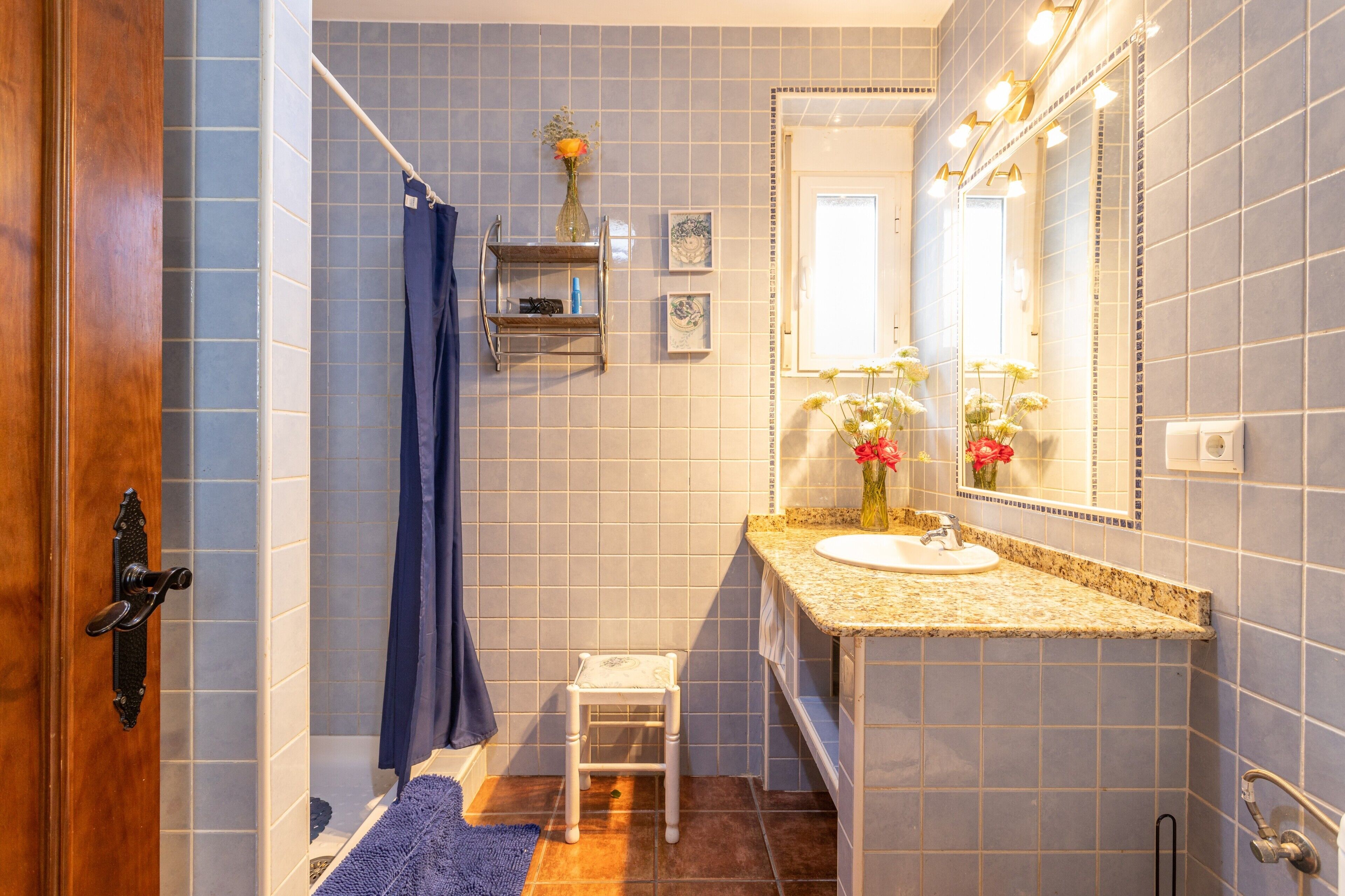 Une salle de bain avec une douche, un lavabo, un miroir et une petite étagère sur laquelle se trouve un vase.