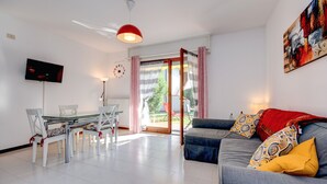 A modern living room with a glass dining table, a grey sofa with patterned cushions, a red pendant light, and a TV mounted on the wall. - Great Location with Charming Patio - Prato Verde - Girasole (San Felice del Benaco)