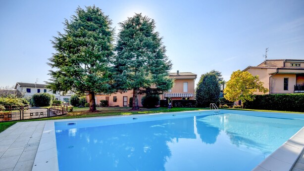 A swimming pool with a clear blue surface, surrounded by a paved deck. There are trees and buildings in the background. - Great Location with Charming Patio - Prato Verde - Girasole (San Felice del Benaco)