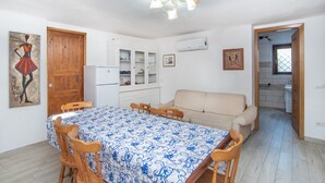 Un espacio habitable compacto con una mesa de comedor, sillas de madera, un sofá, un ventilador de techo y una cocina visible a través de un vano abierto.